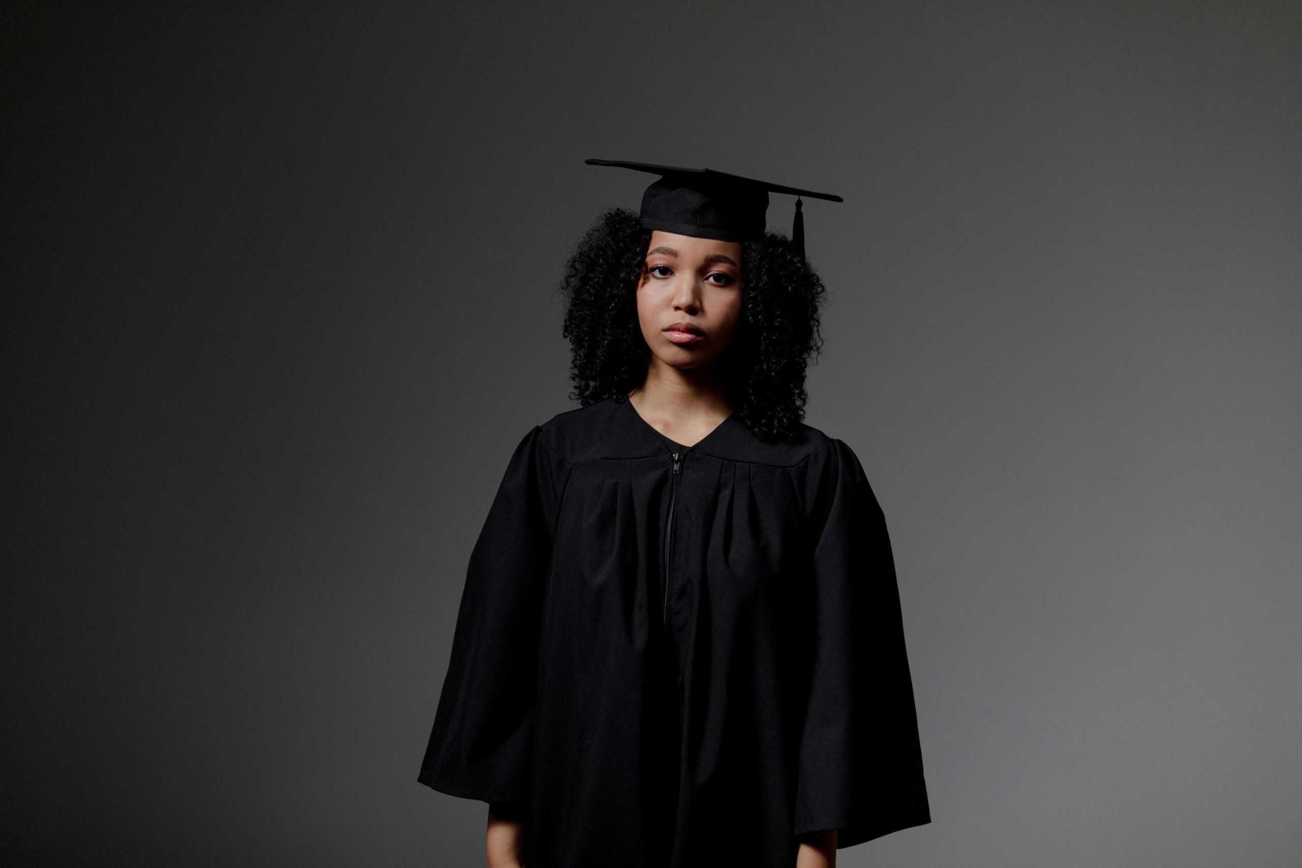 Portrait of a young female graduate in a cap and gown, with a neutral expression against a dark background.