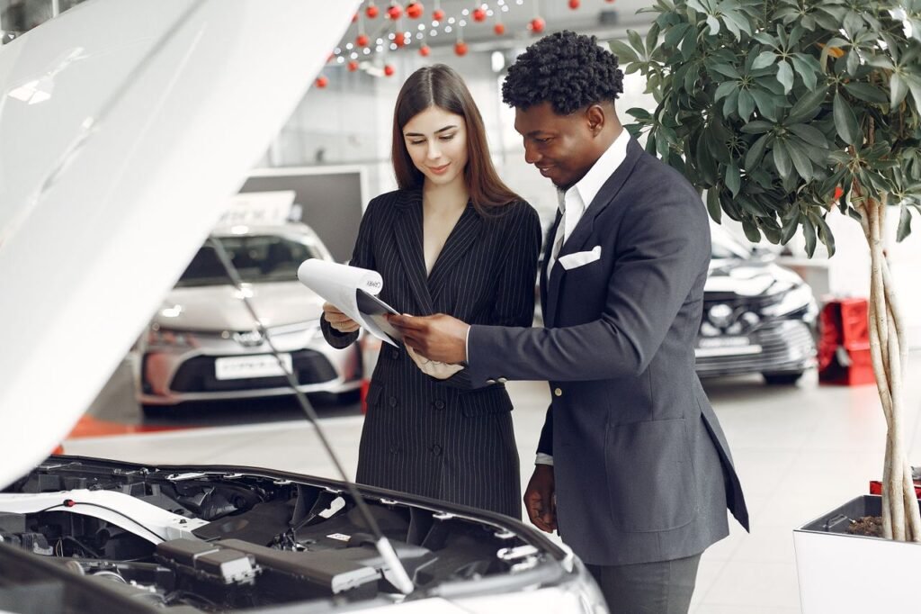 CAR LOAN USA REVIEW Confident smiling successful black businessman in classy formal suit standing near open motor hood in car showroom and discussing car characteristics with gorgeous female dealer