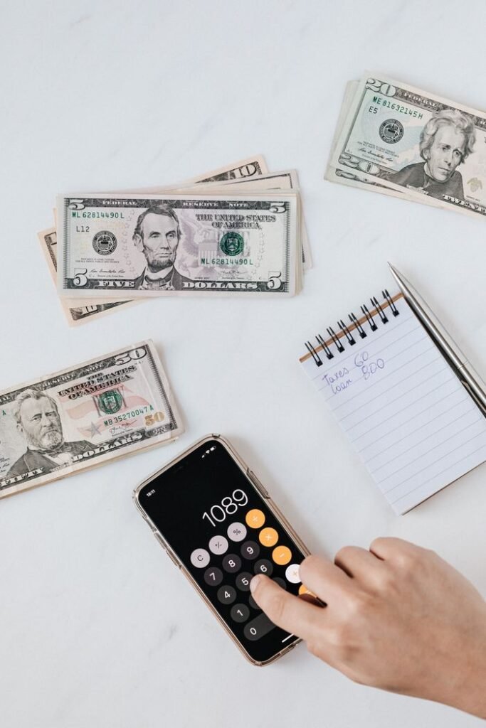 PERSONAL LOANS From above of crop unrecognizable person using calculator app on cellphone while calculating total amount of money sitting at table with different piles of American dollars and notebook with notes